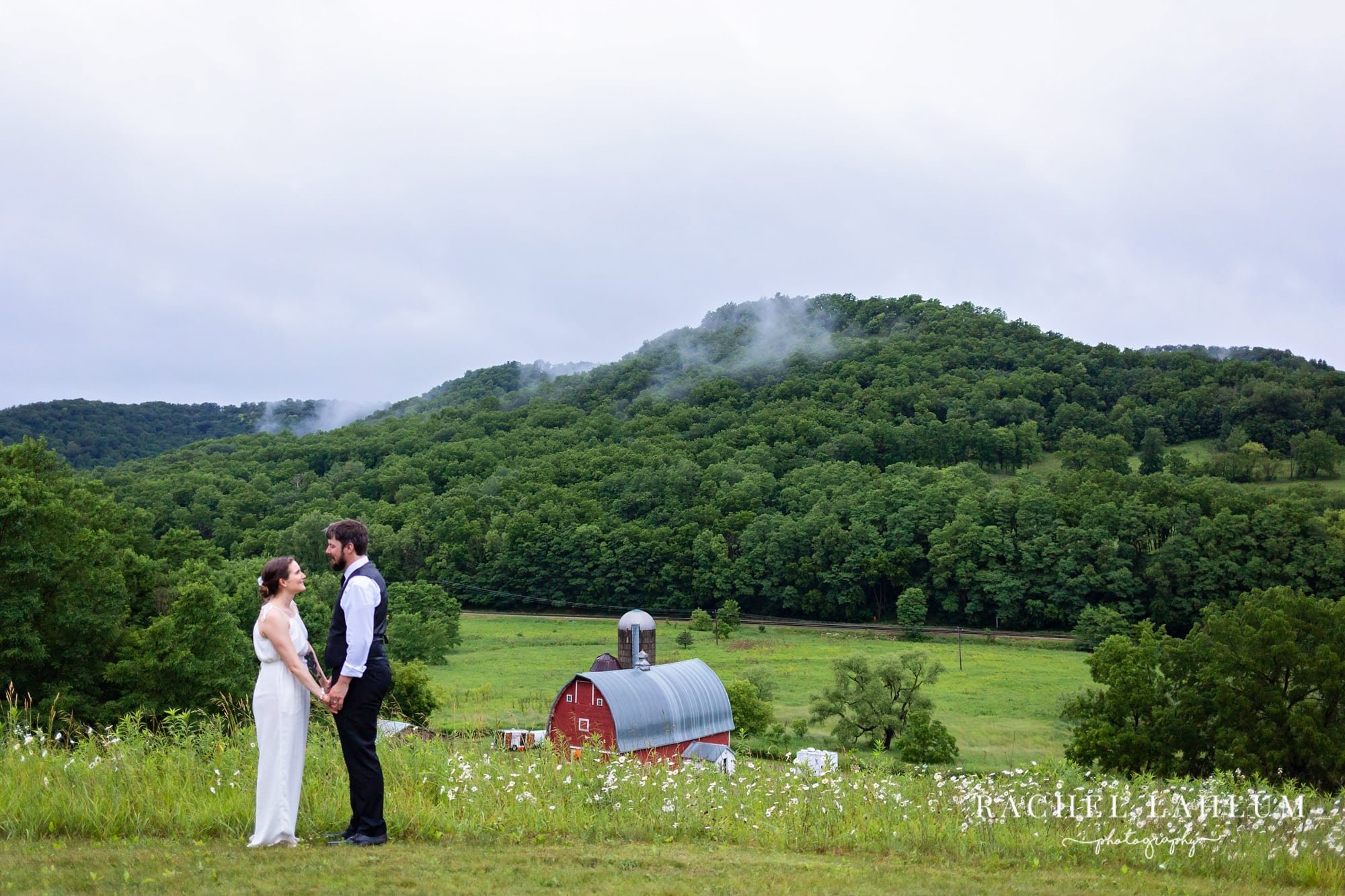 Newly-wed couple hold hands outside on a hill overlooking Cedar Creek Barn and the hills of Winona, Minnesota. 