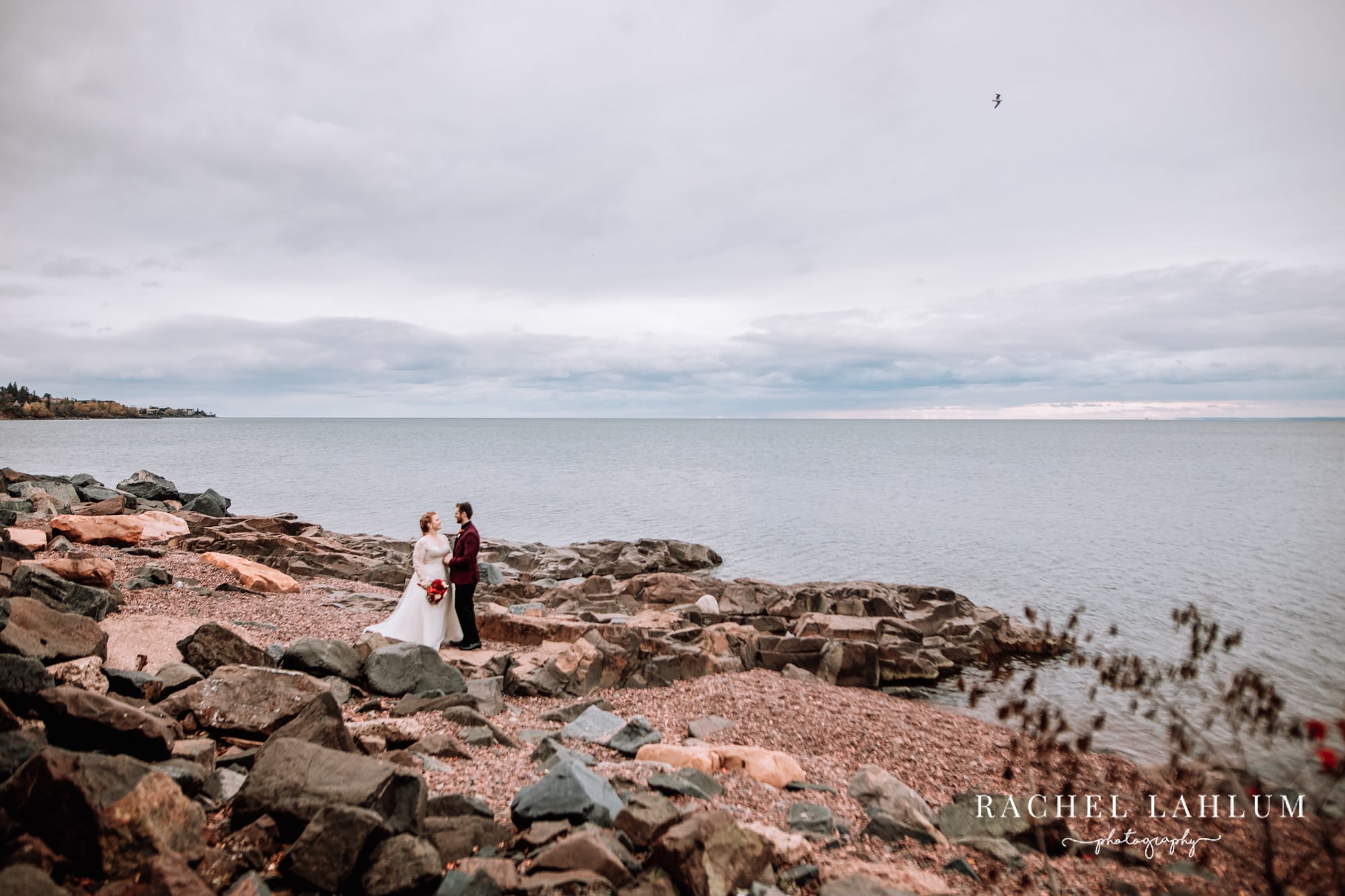 Bride and groom hold each other on a rock beach overlooking Lake Superior in Duluth, Minnesota.