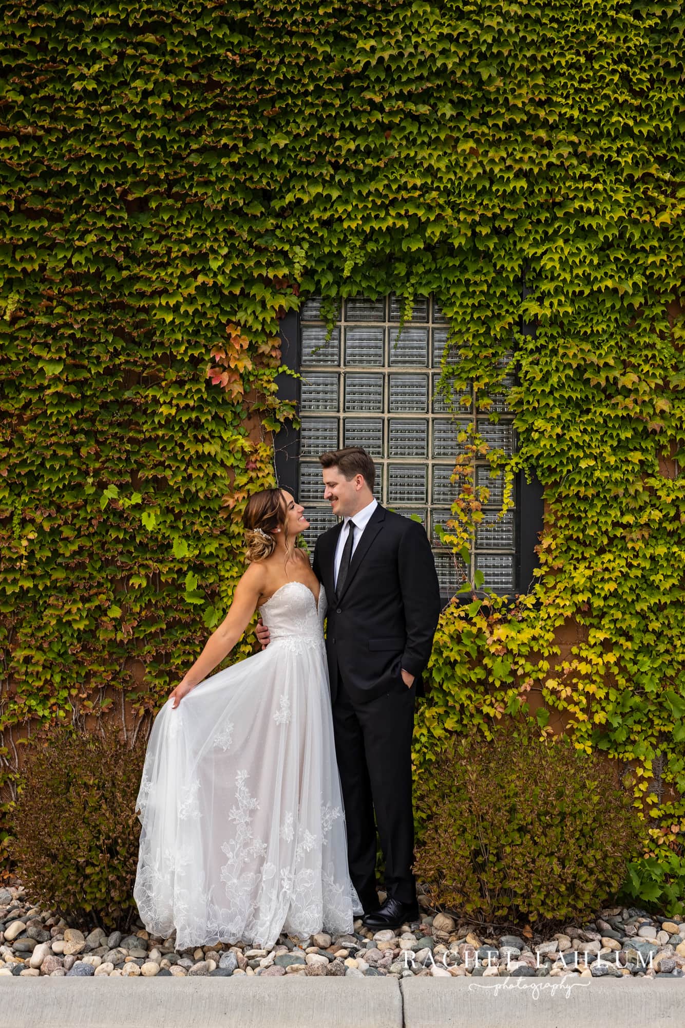 Bride and groom smile at each other during wedding photography, while standing in front of a wall covered in ivy at the Boathouse in Alexandria, Minnesota.