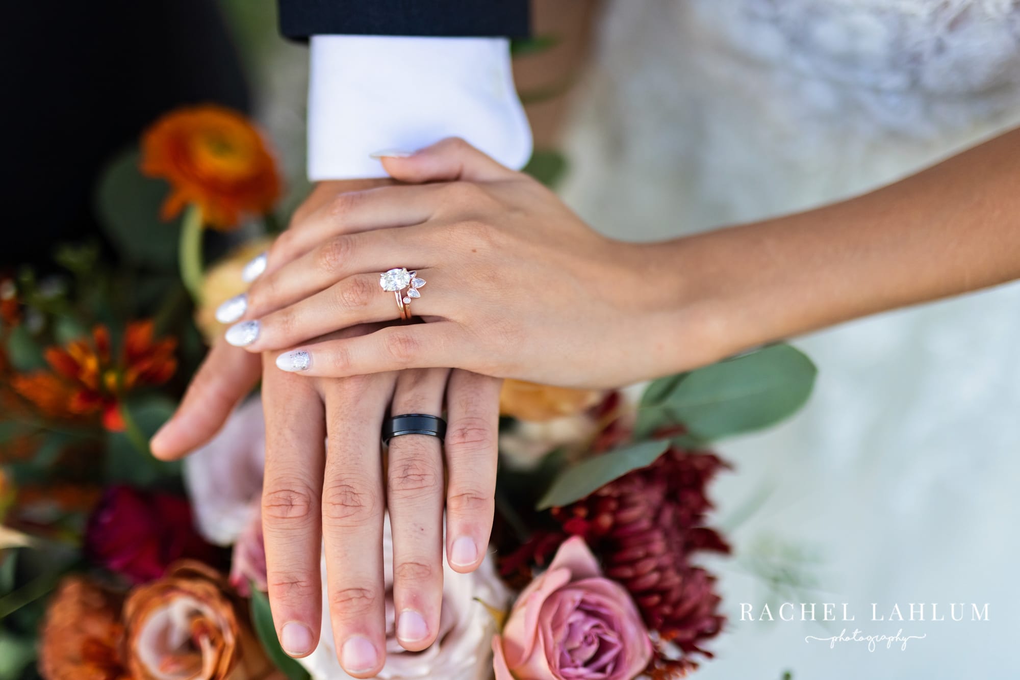 Bride’s hand lies on top of grooms hand showing wedding rings over the bouquet.