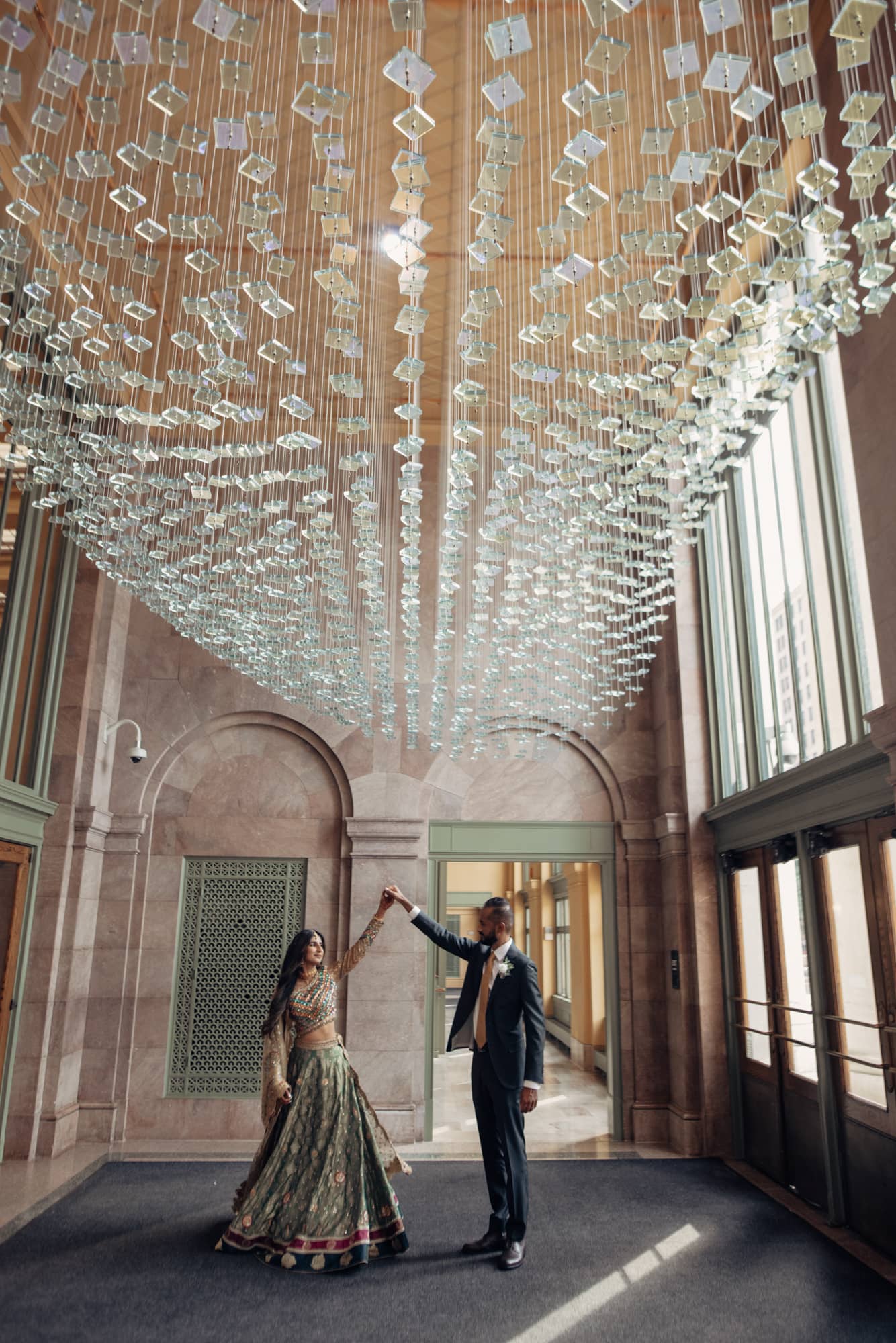 Groom twirls bride under glass ceiling fixture inside the lobby of Saint Paul Union Depot. 