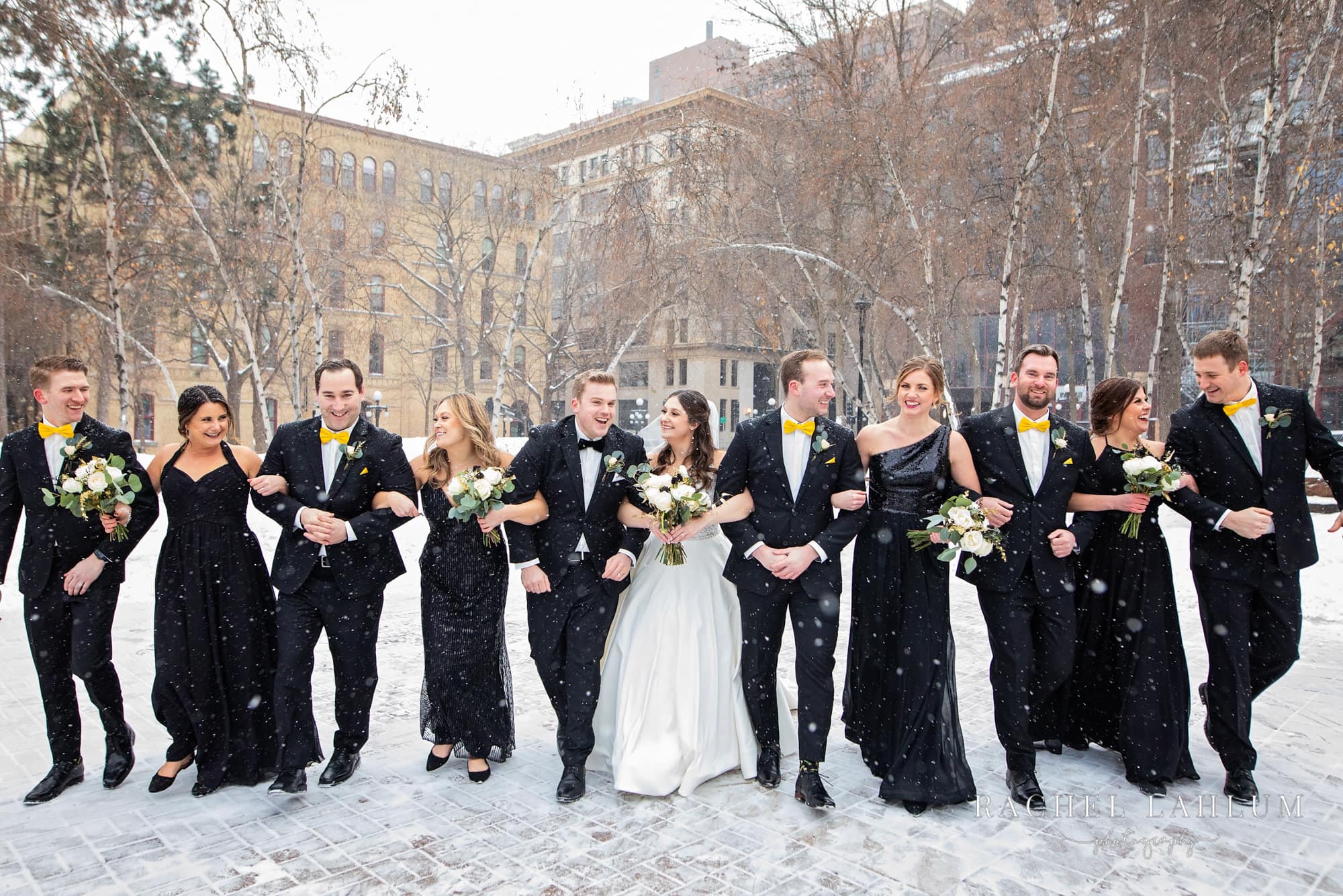 Bridal party lock arms and walk forward in snowy winter wedding at Mears Park in St. Paul, MN.