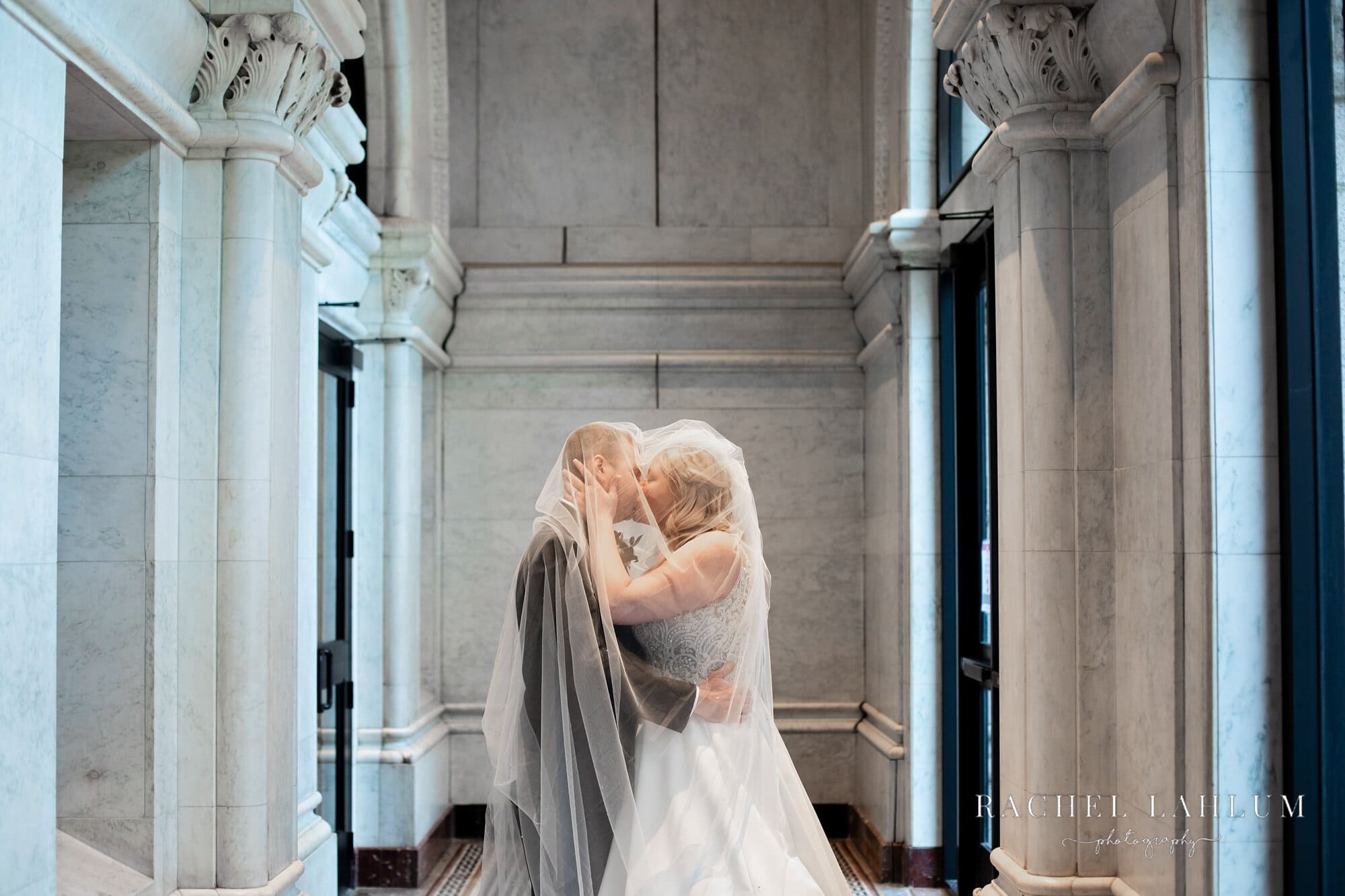 Bride and groom kiss under the bride’s veil in marble entryway of the Landmark Center. 