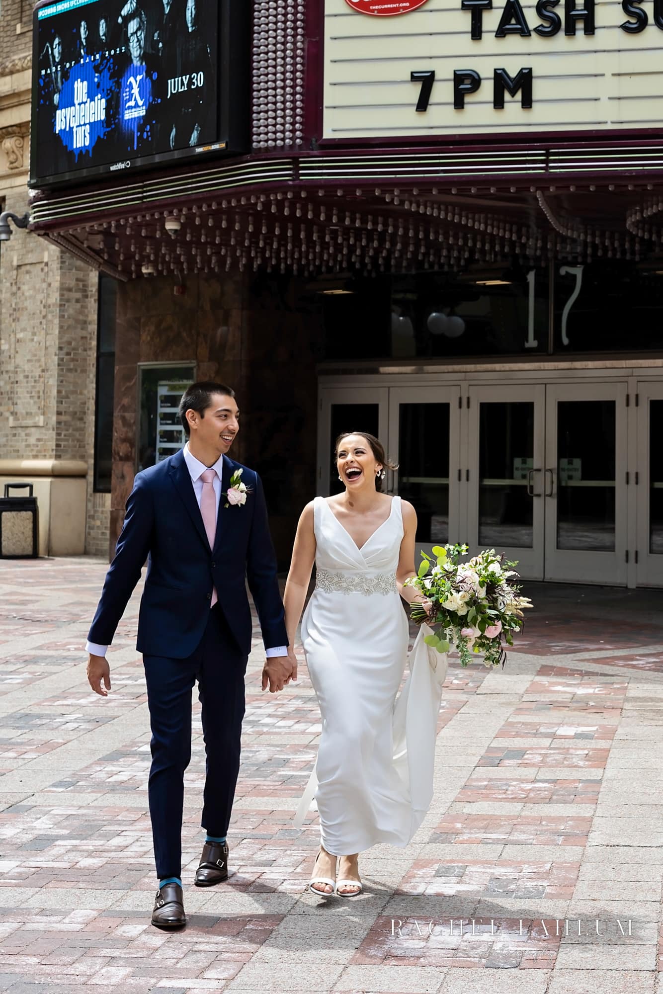 Bride and groom hold hands and laugh while walking away from historic theater by St. Paul’s Seventh Place Plaza.