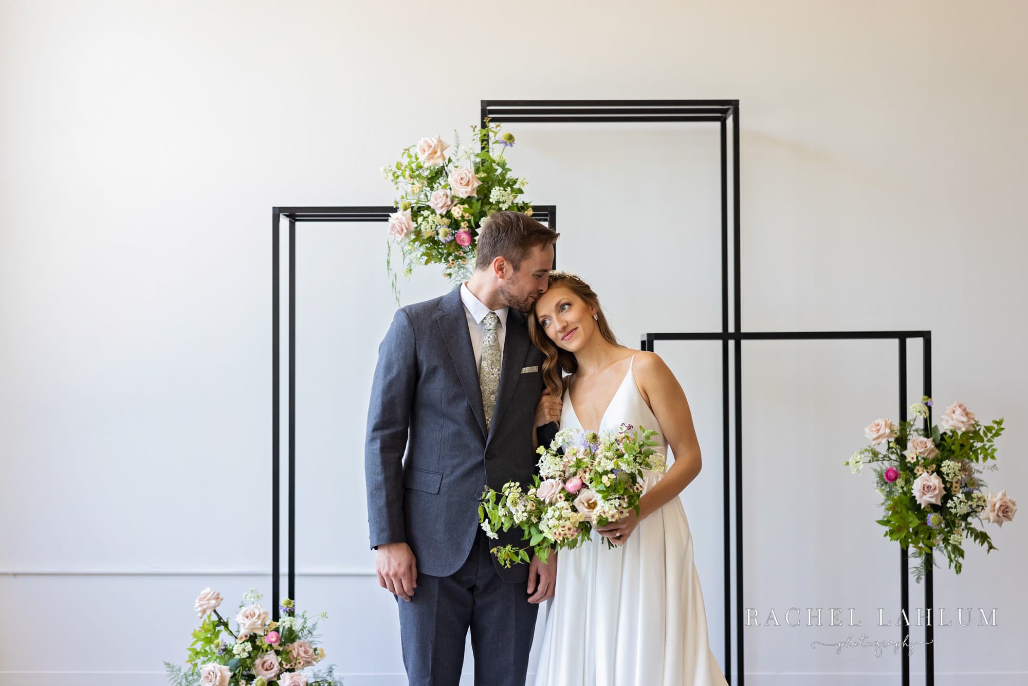 Groom kisses bride on head in front of metal fixture with floral arrangements at Northeast Minneapolis wedding space. 