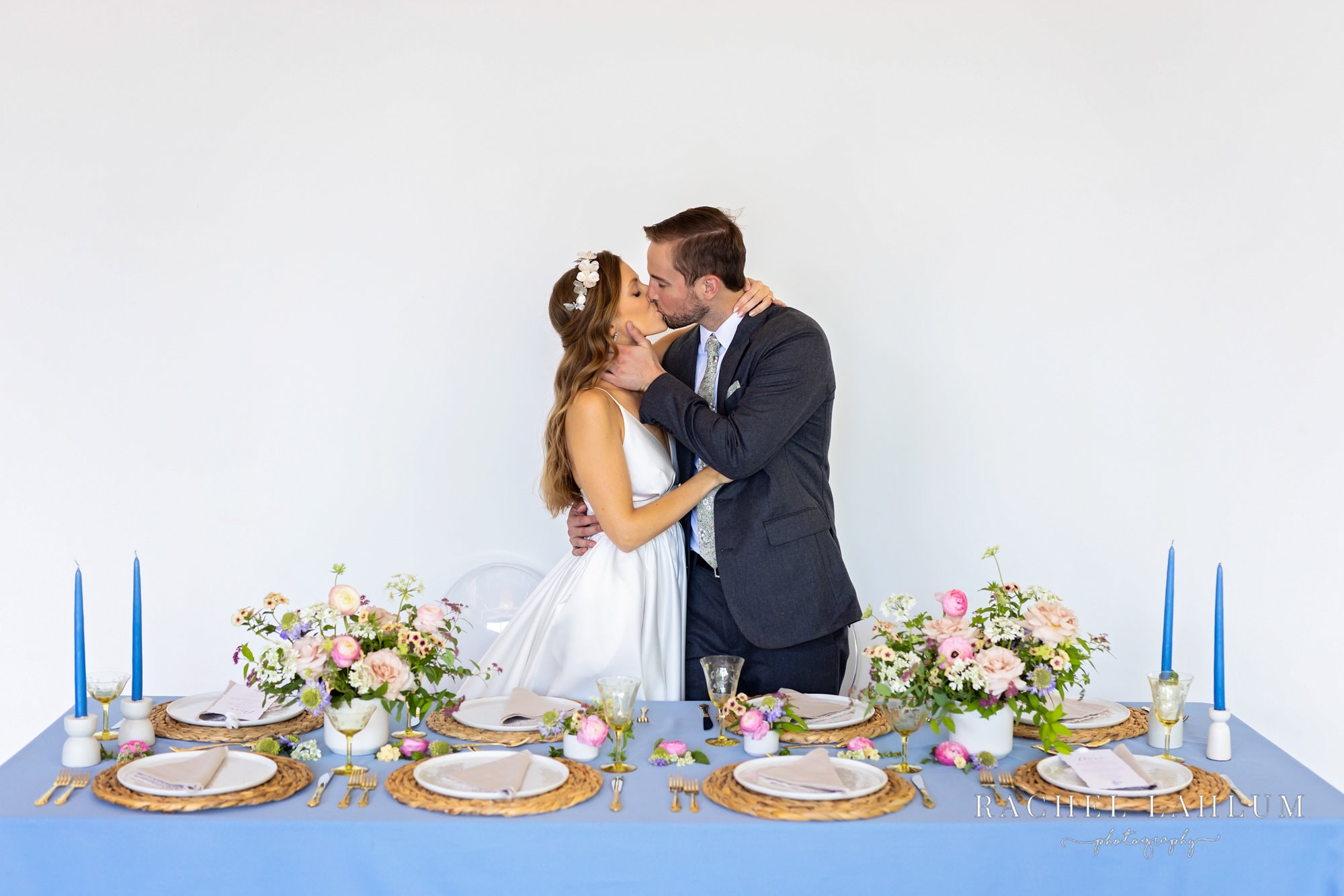 Bride and groom kiss behind spring-like table place settings at stylized wedding shoot. 