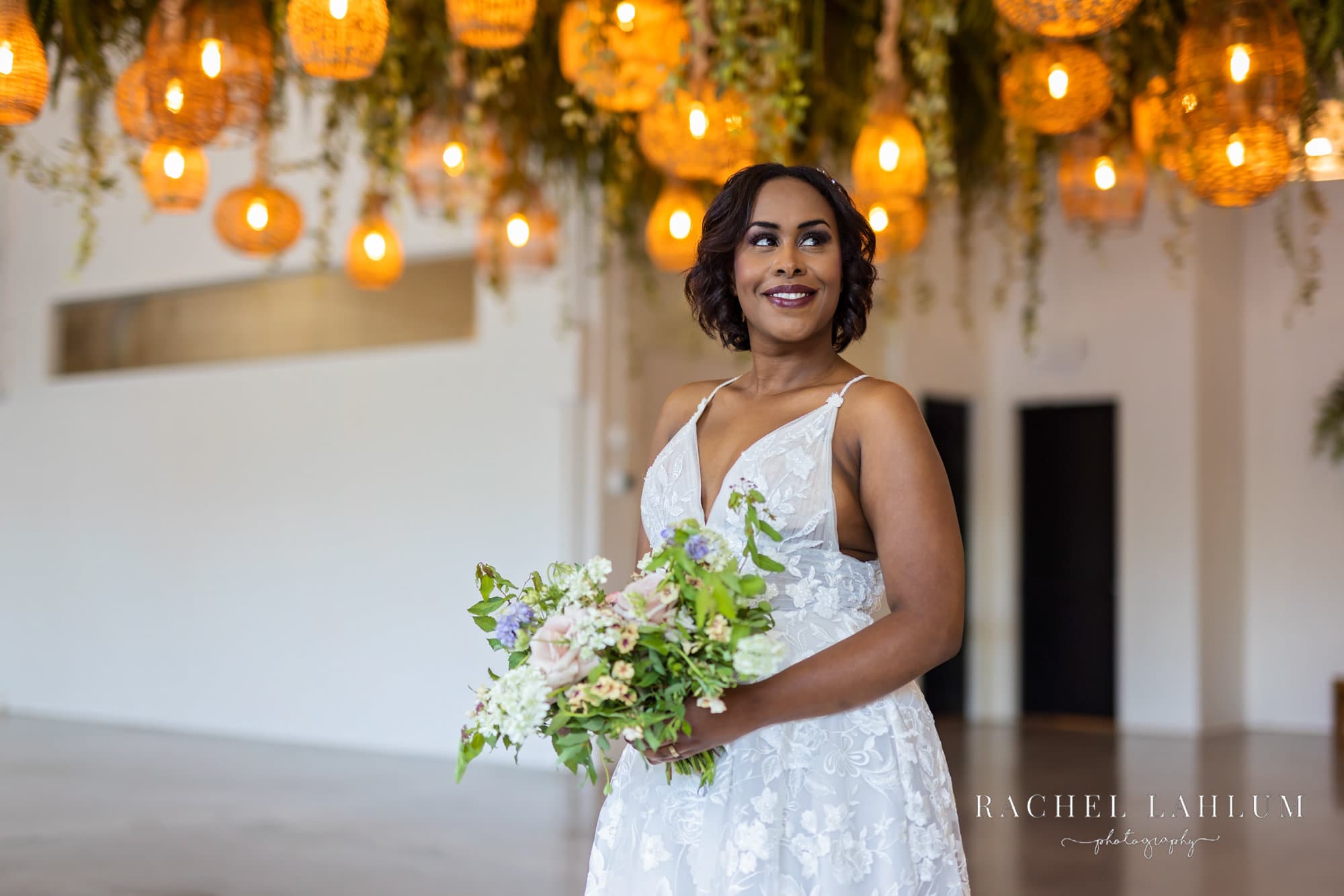 Bride poses its bouquet under greenery and lighting fixture during stylized wedding photo shoot at Urban Daisy Event Space.