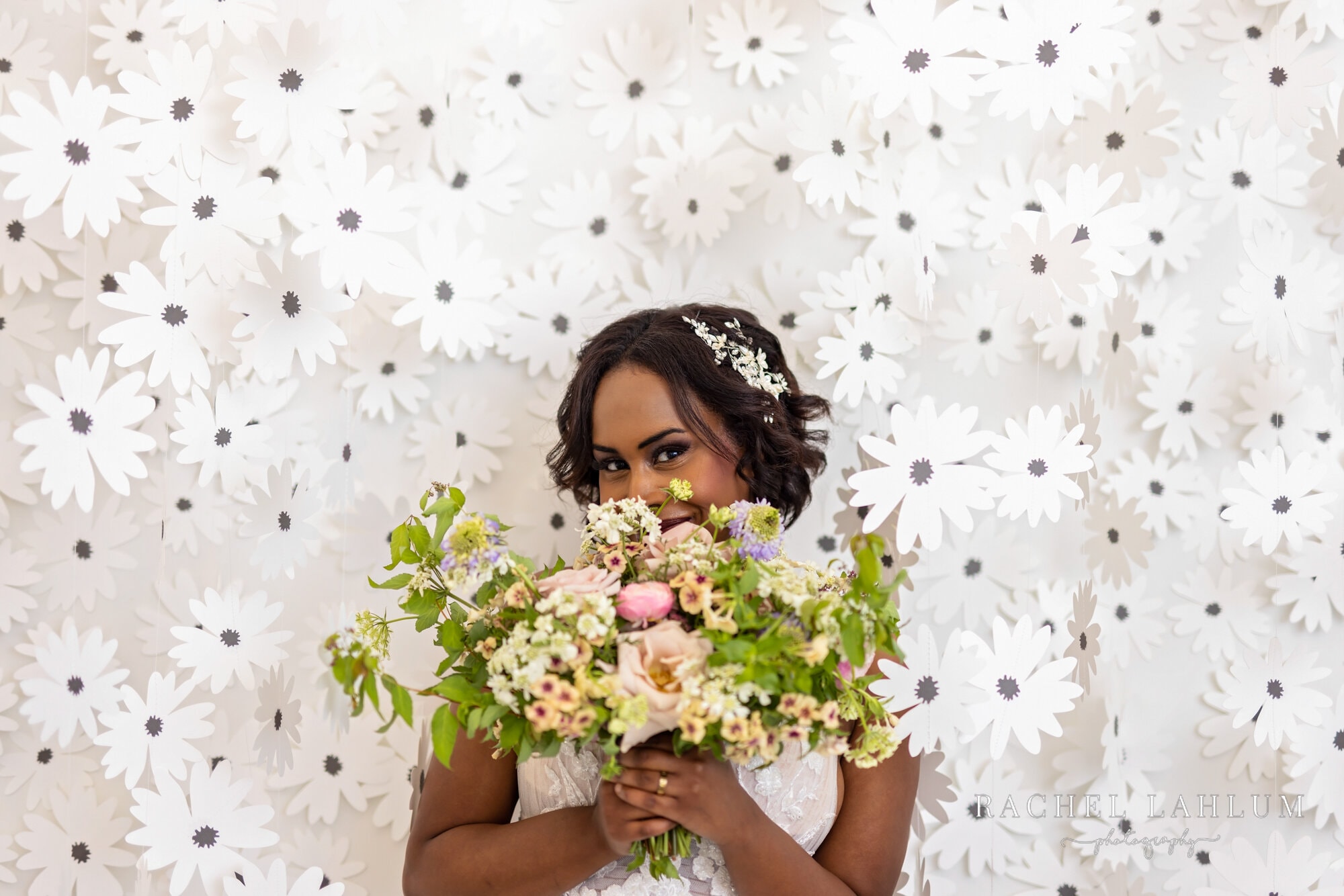 Bride smiles behind bouquet in front of flower-decorated wall at Urban Daisy Event Space in Northeast Minneapolis. 
