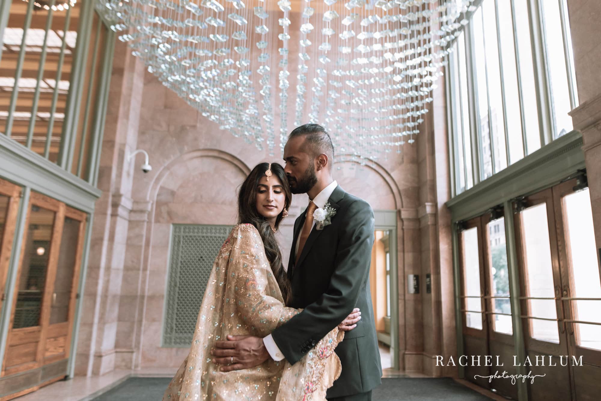 Bride and groom embrace inside the lobby of the Intercontinental with glass ceiling decorations above. 