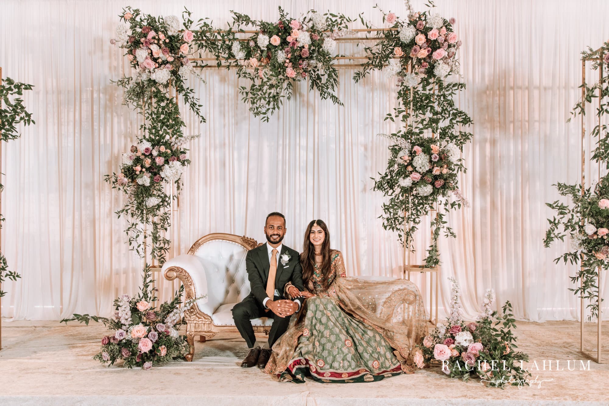 Bride and groom pose for a portrait beneath floral decorations surrounding a white and gold couch. 