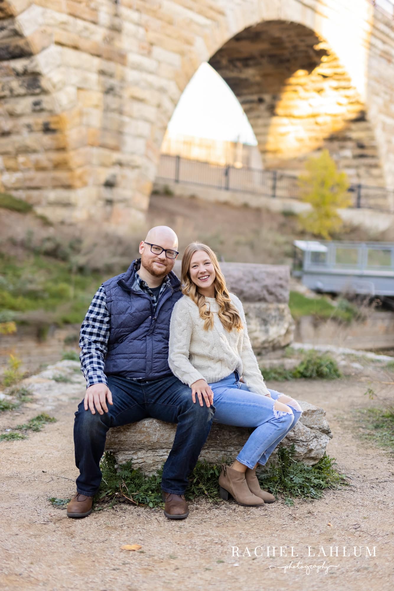 New engaged couple pose while sitting on a rock under the Minneapolis Stone Arch Bridge.