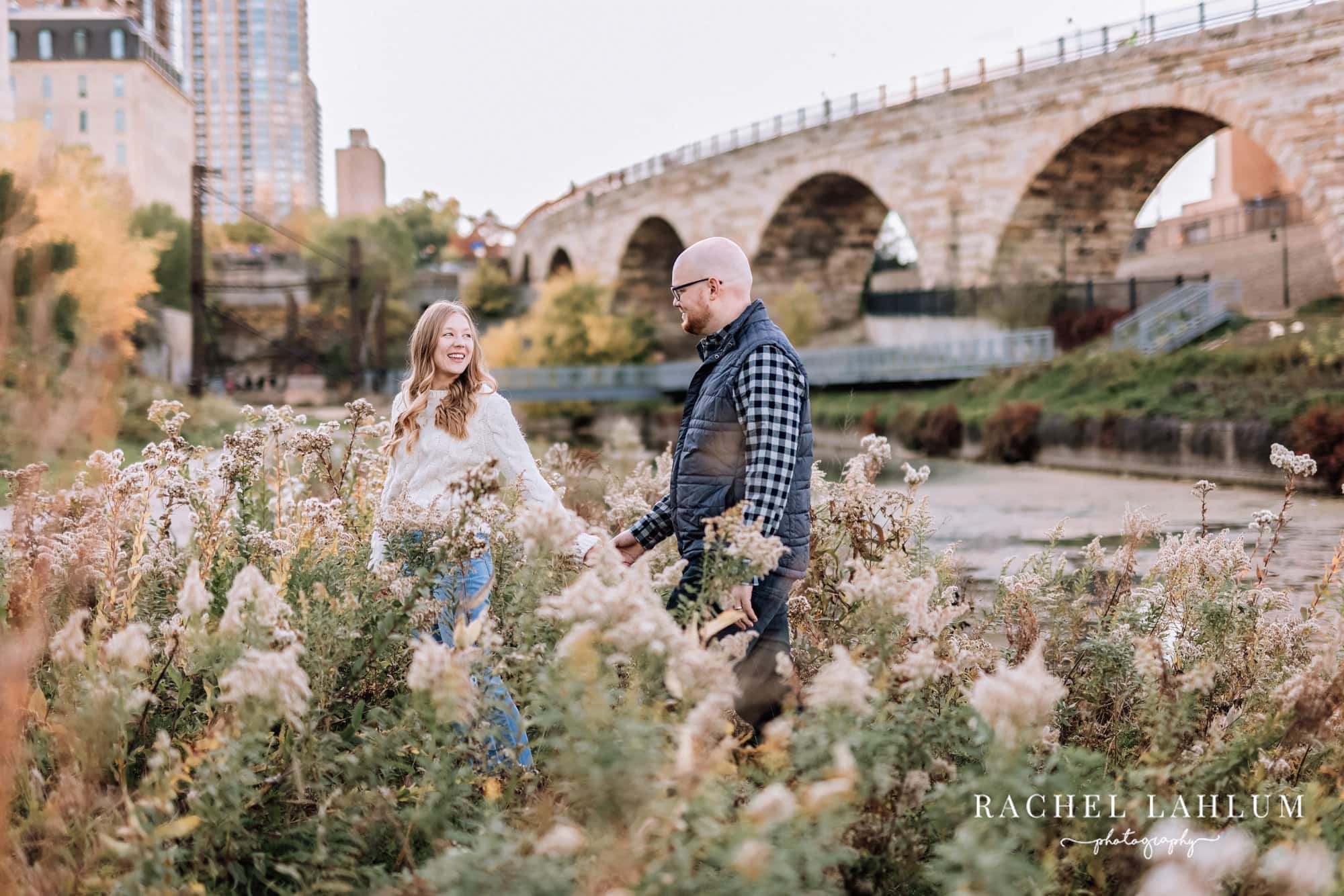 Woman leads her fiancé through garden by the Stone Arch Bridge in Minneapolis.