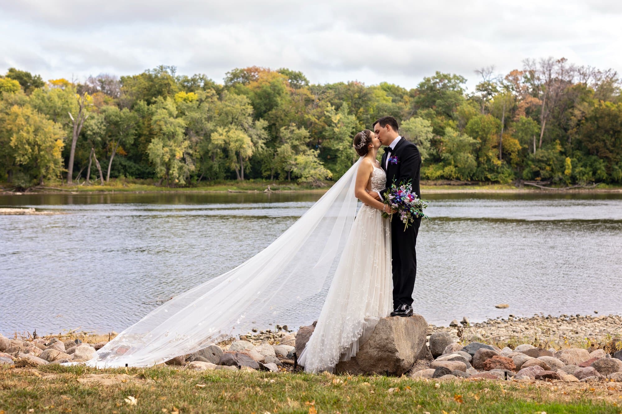 A landscape wedding portrait of a bride and groom kissing in front of the Missisippi river with rocks and a beautiful treeline behind them, the bride's veil elegantly stretching across half the photo, in Leopold's Mississippi Gardens, NE Minneapolis