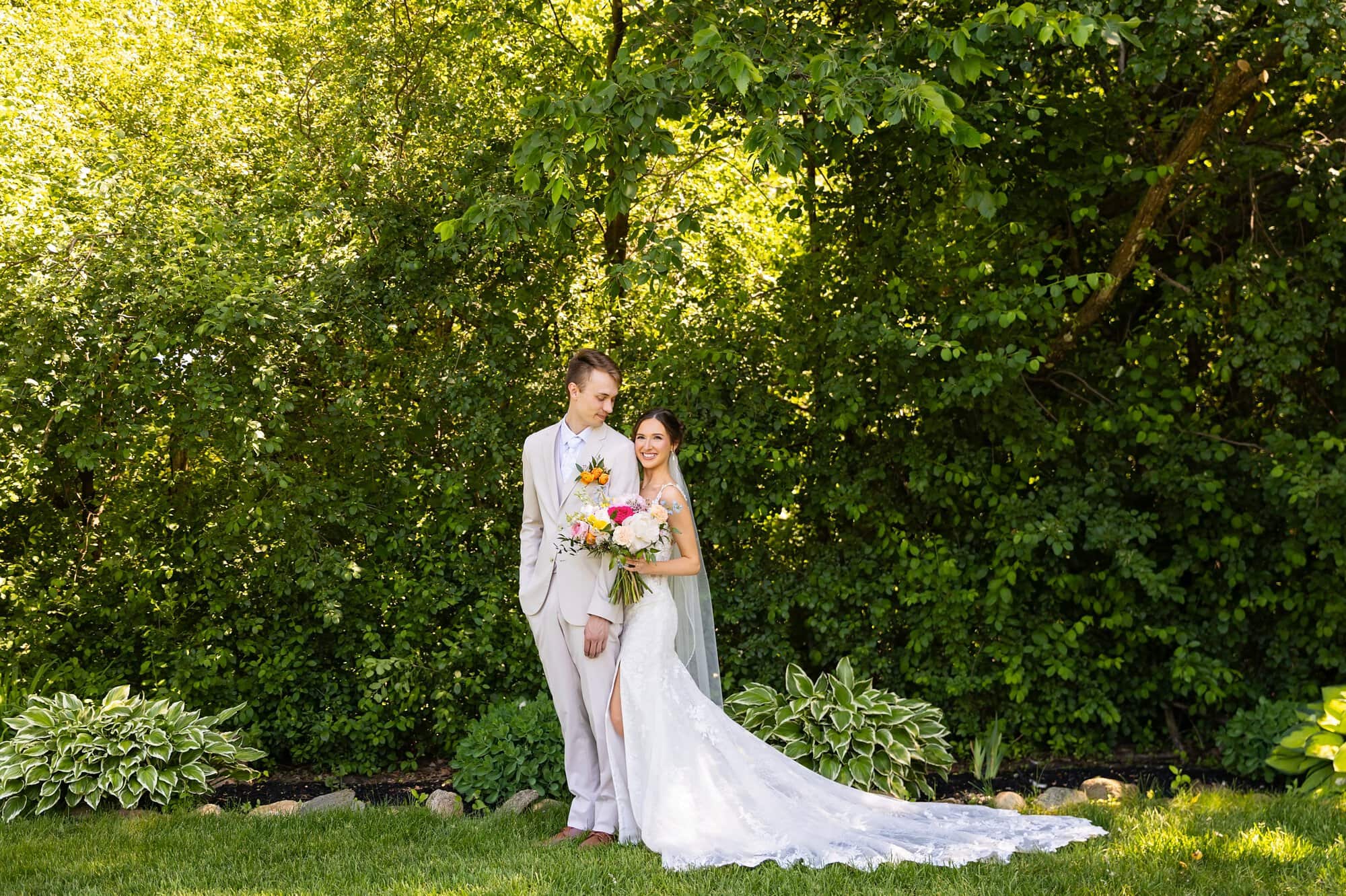 Wide landscape portait of the wedding couple, bride and groom, surrounded by lush, green foliage at Ridgetop Events. Groom looking at the fiancee and bride smiling at the camera.