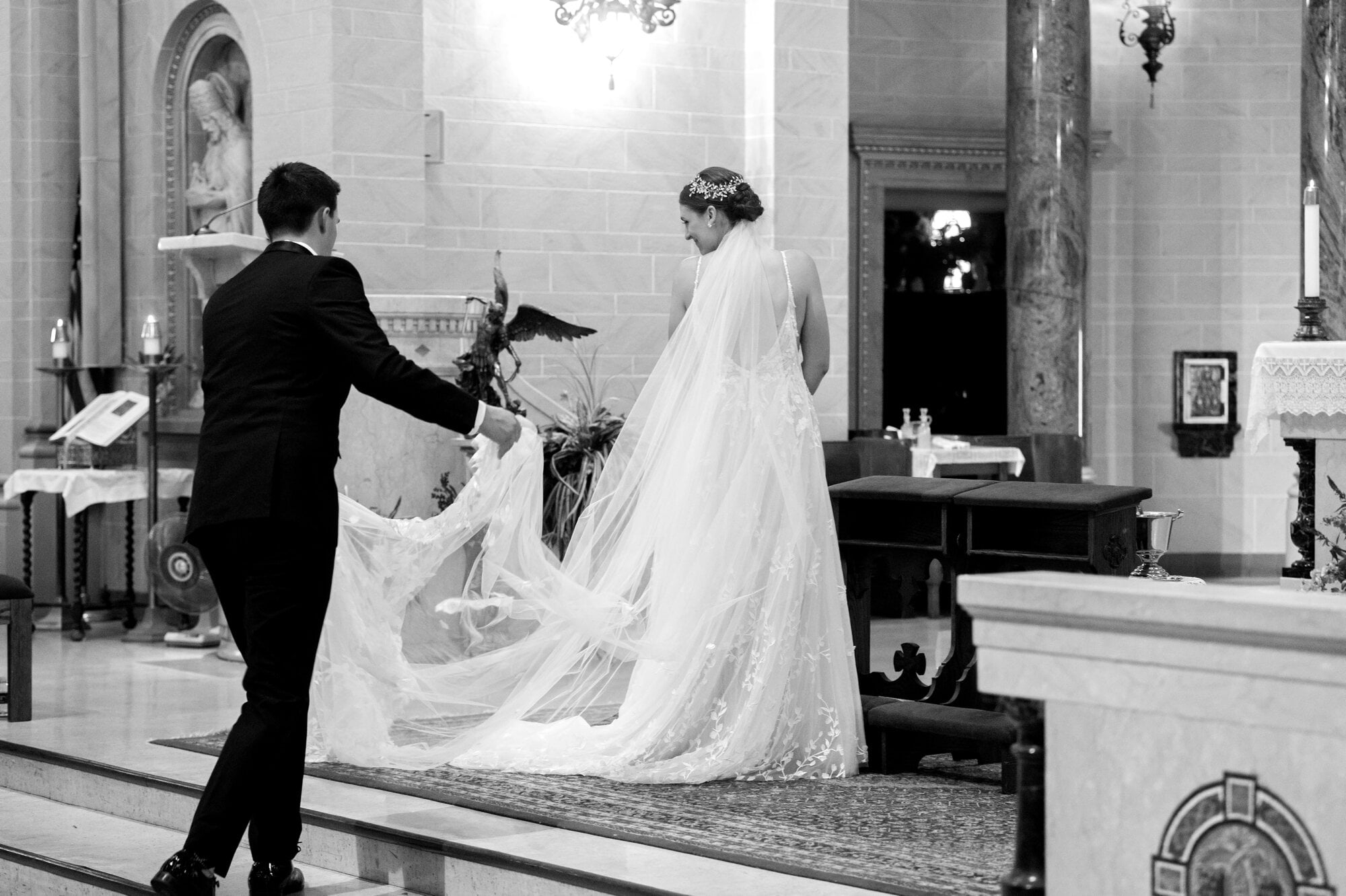 Candid black and white portait of the groom fixing the bride's wedding veil train inside of the Holy Cross Catholic Church, NE Minnepolis