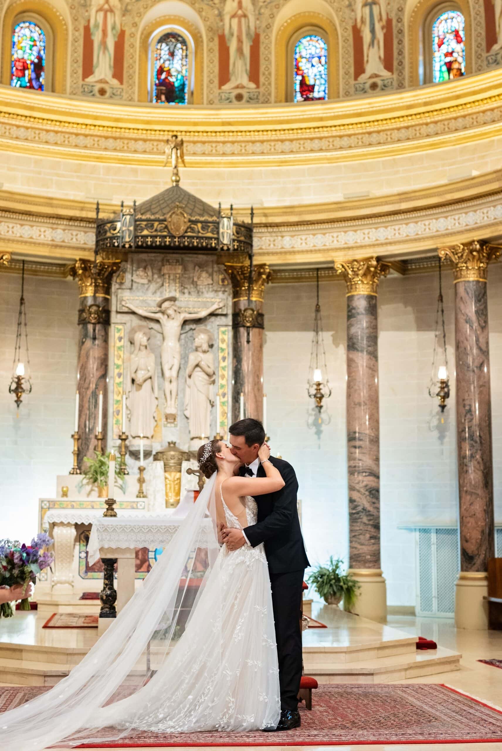 A stunning view of the architecture of the building as the bride and groom kiss in front of the altar, crucifixion scene and gilded marble columns at the Holy Cross Catholic Church in NE Minneapolis