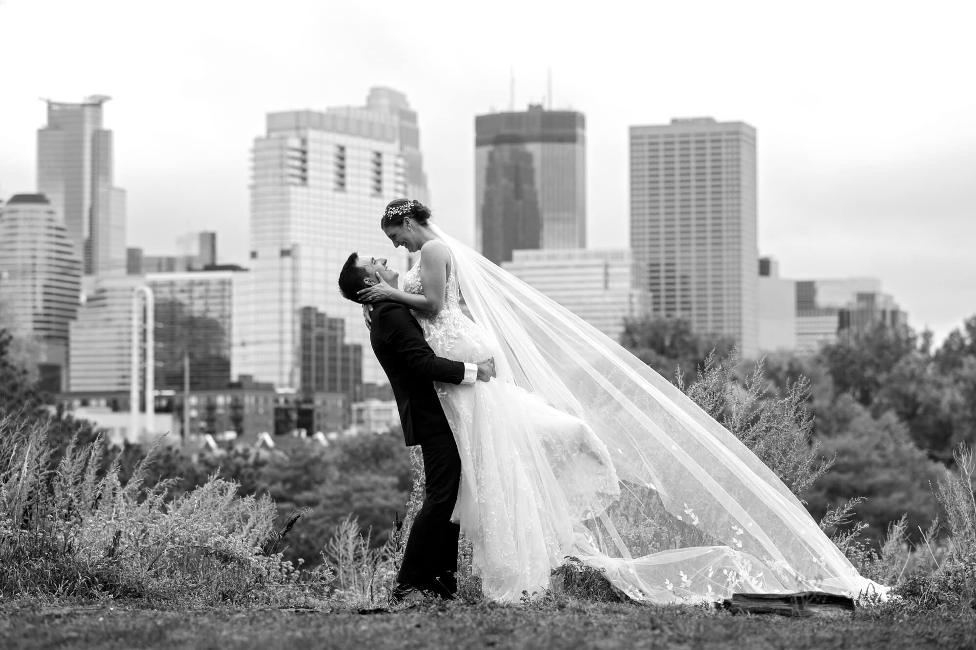 Groom lifts up his bride in a black and white wedding portait highlighting the NE Minneapolis skyline behind them with the bridal veil floating out behind