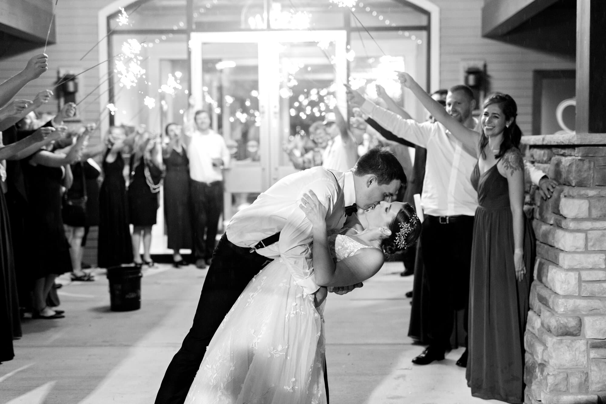 Black and white portrait of wedding guests framing the walkway with raised sparklers as the bride and groom kiss outside the reception at Leopold's Mississippi Gardens, NE Minneapolis