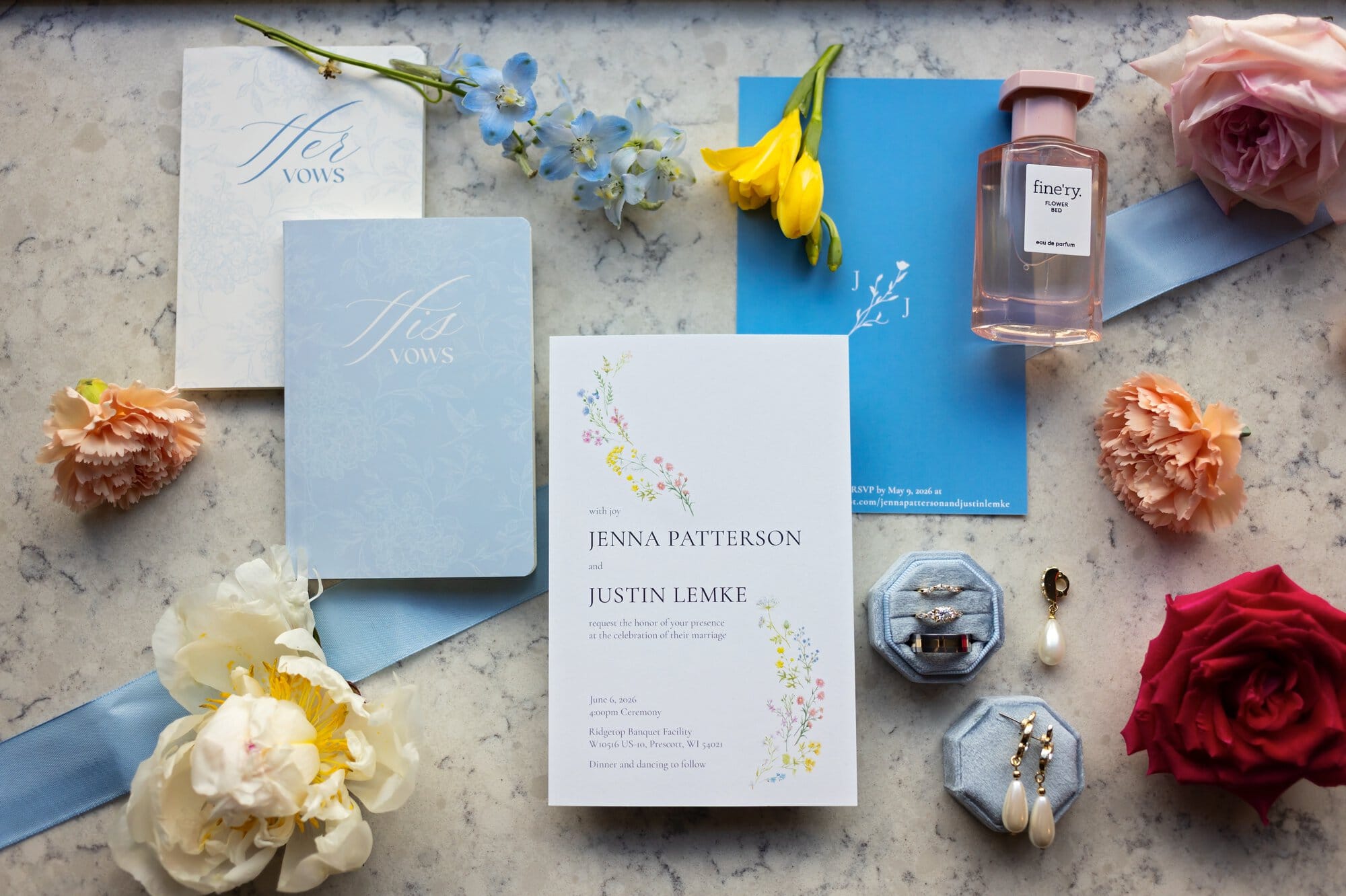 Flat lay of Jenna and Justin's wedding invitation suite featuring custom vow books, wedding rings, pearl earrings, floral details, blue ribbon, and perfume at Ridgetop Events in Prescott, Wisconsin.