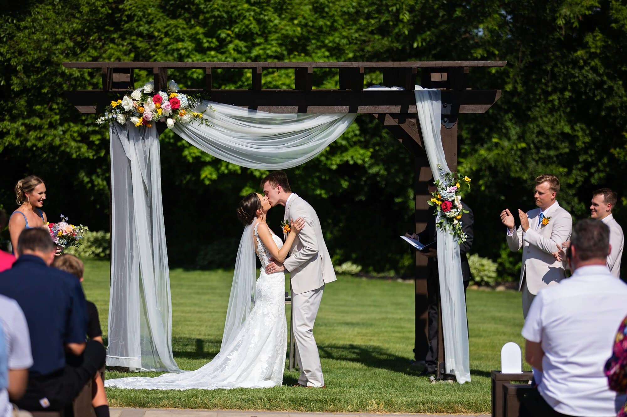 Couple share first kiss as man and wife before the audience under a white tulle decorated pergola with flowers at their Ridgetop Events ceremony in Prescott, WI.