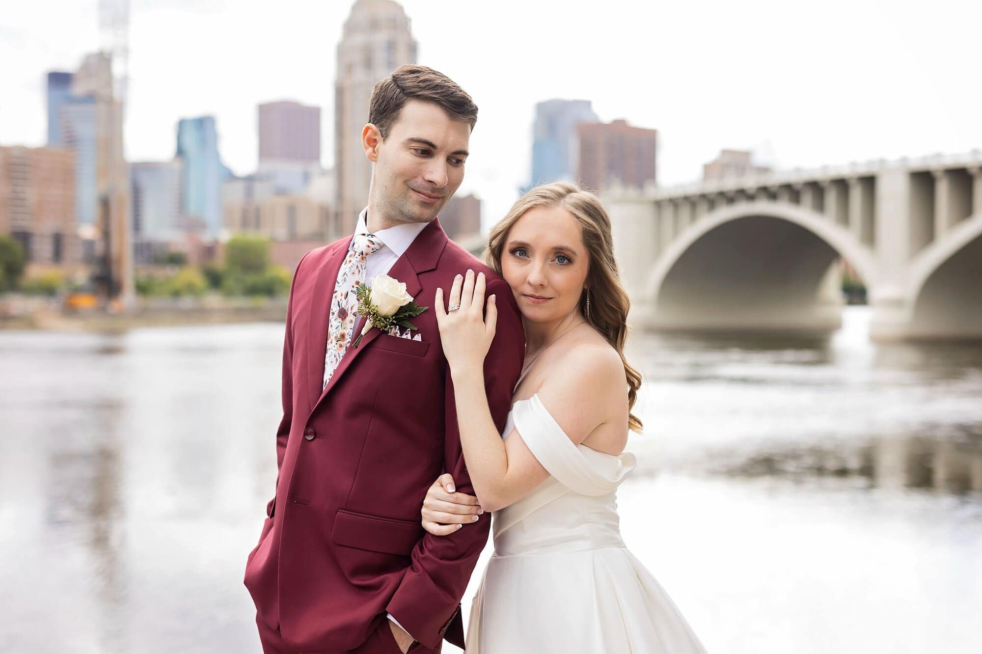 Bride and groom pose for wedding photos on the boardwalk in front of the river and skyline near Nicollet Island Pavilion in Minneapolis Minnesota