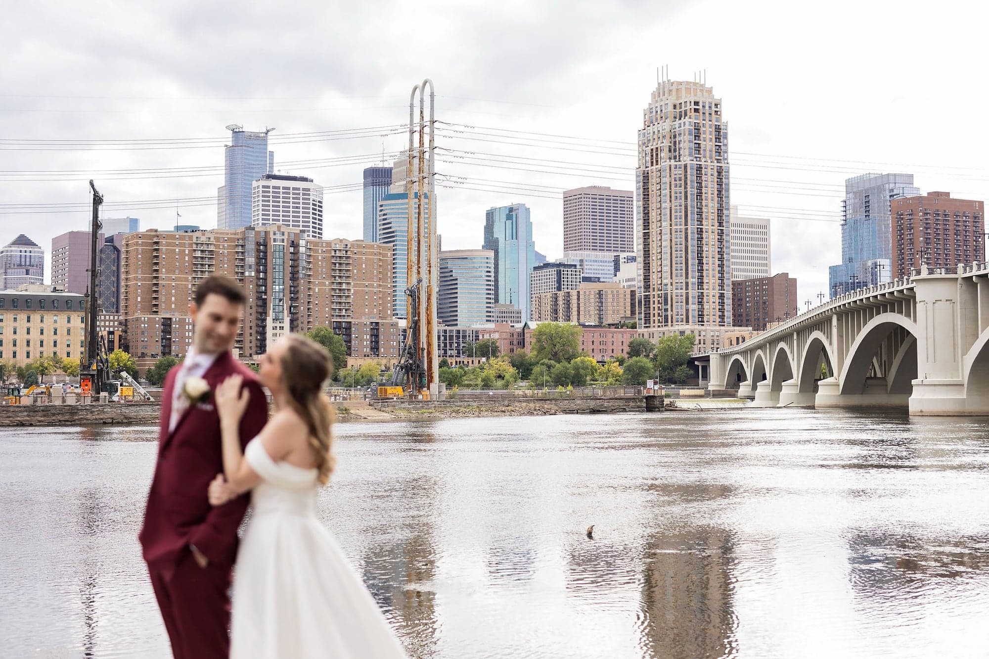 Bride and groom pose for wedding photos in front of the river and skyline near Nicollet Island Pavilion in Minneapolis Minnesota