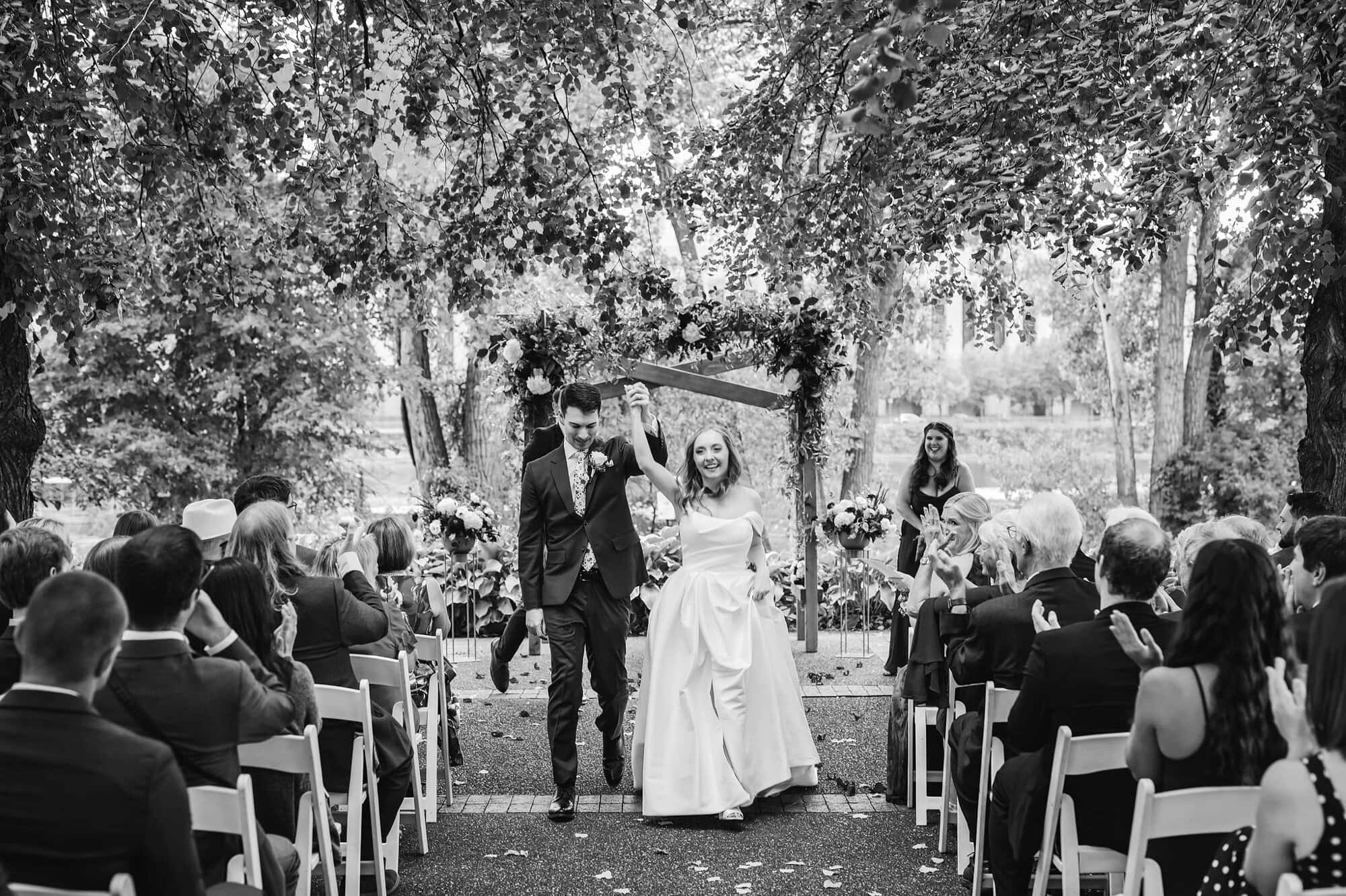 Bride and groom walk down the aisle together in a black and white photo at a wedding ceremony on the Nicollet Island Pavilion patio in Minneapolis Minnesota