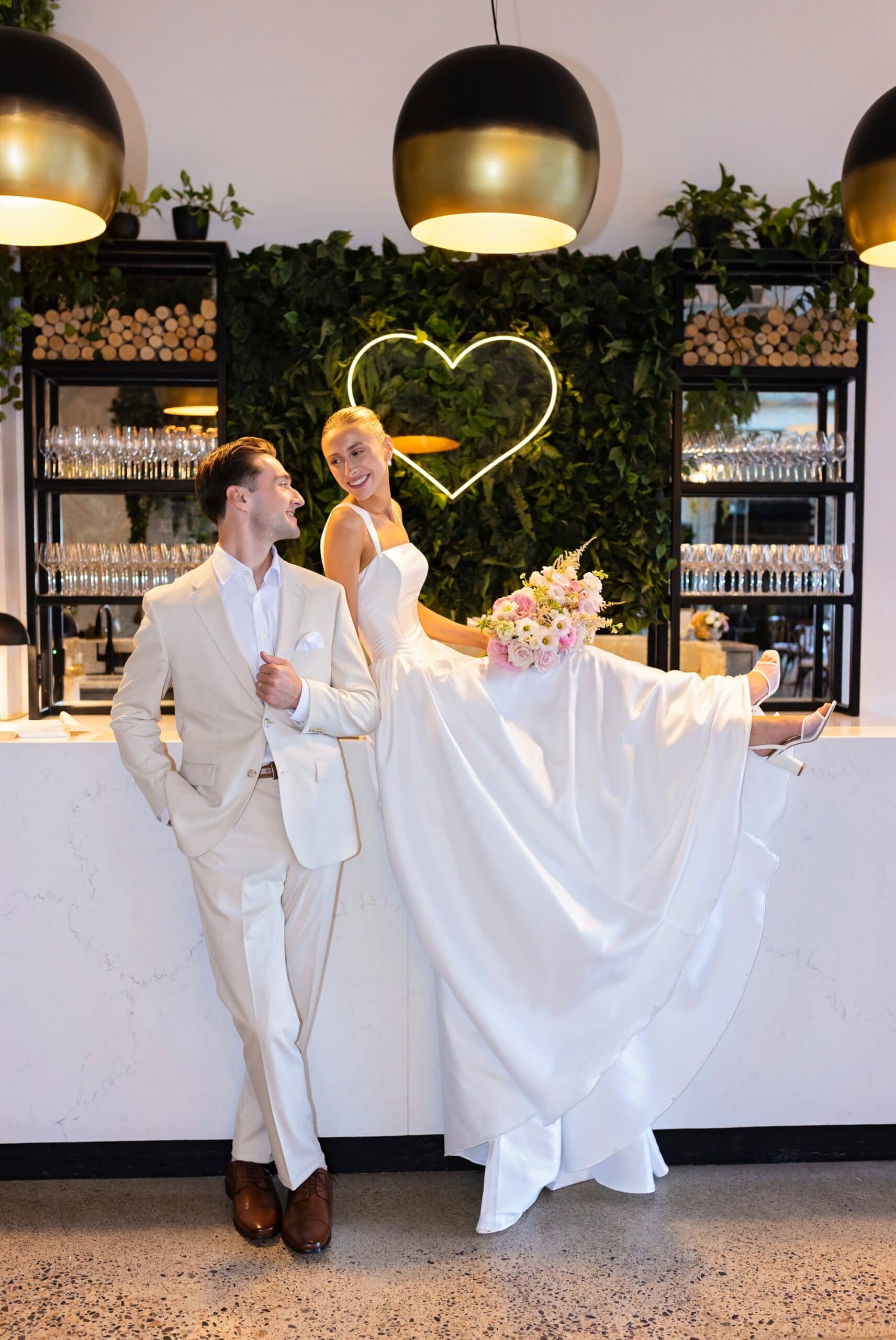 Bride and groom pose with the bride sitting on top of the bar and a lit-up heart set on top of a leafy background in the background during couple photos at the Urban Daisy in Minneapolis, Minnesota