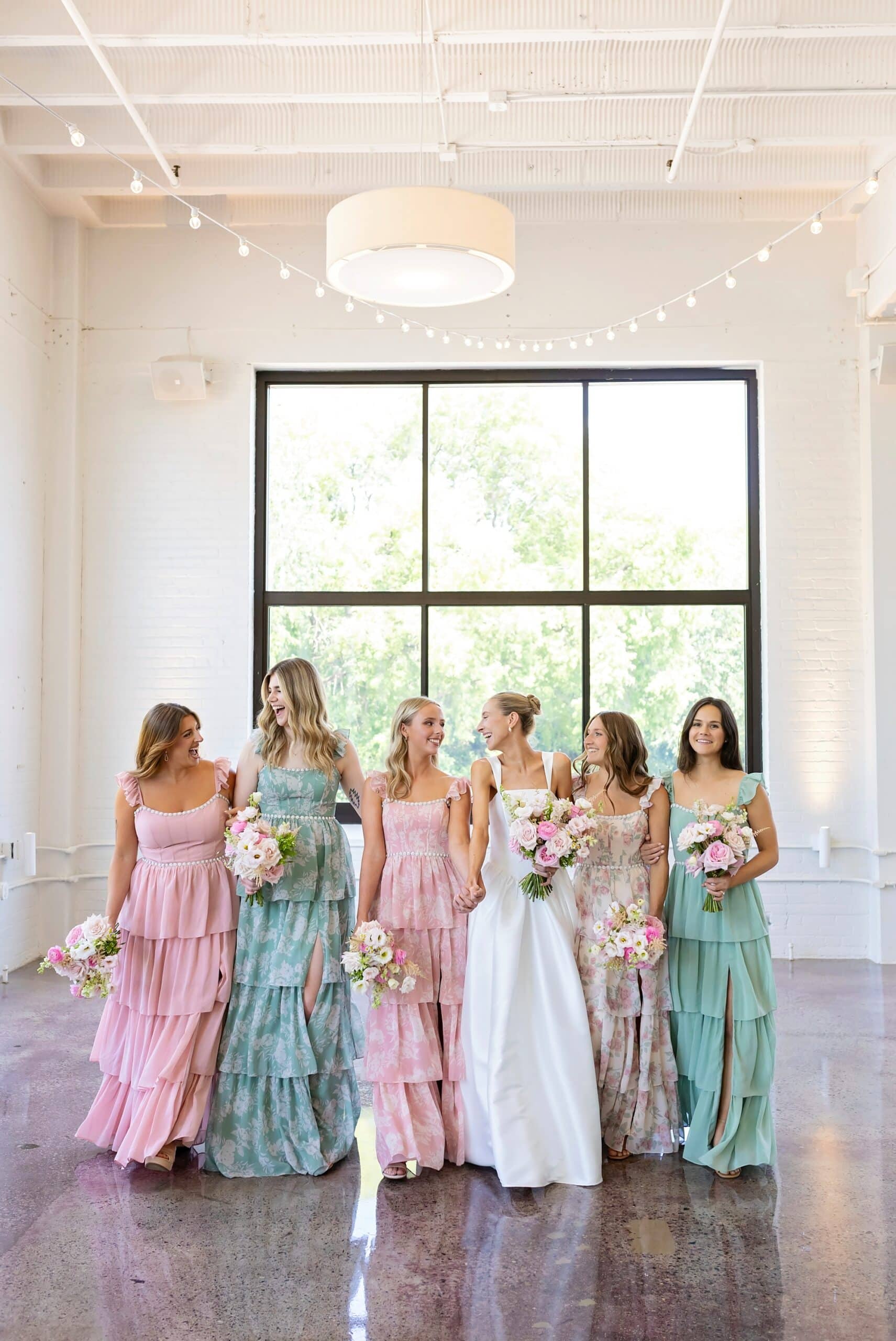 The bride and her bridesmaids link arms in a line showing off their floral pink and green dresses and beautiful bouquets during pre-ceremony wedding photos at the Urban Daisy in Minneapolis, Minnesota